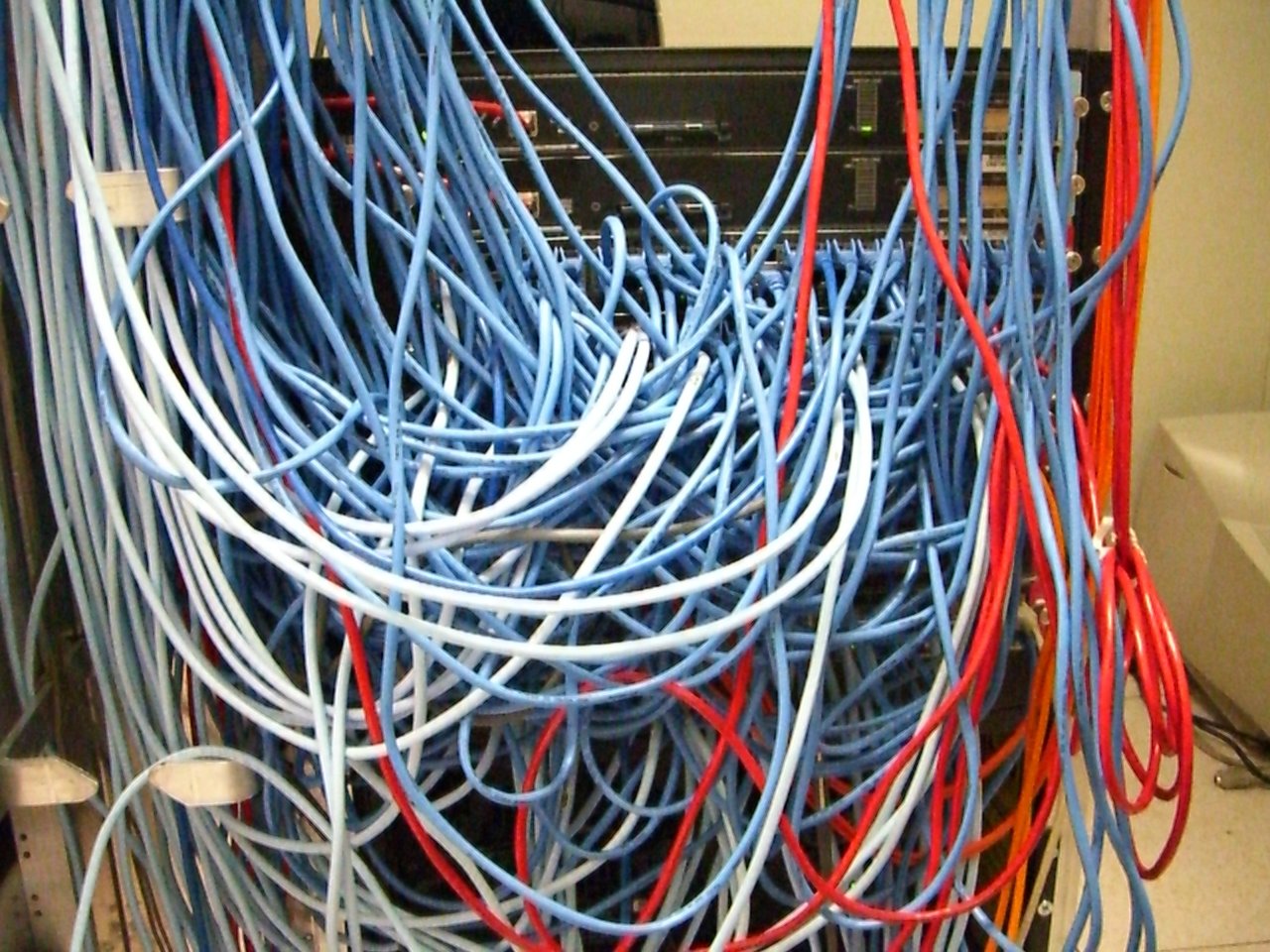 Photo of a tangled mass of blue, white, and red network cables running into a server rack, representing the internet-facing network infrastructure exposed by the MikroTrick attack chain